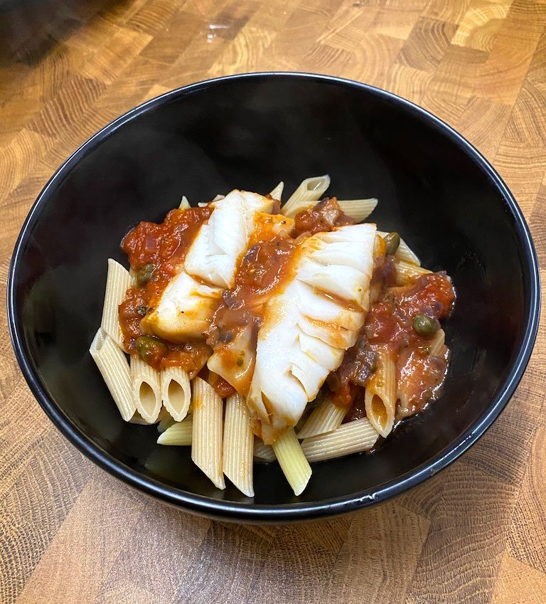 Tuscan baked fish with tomato sauce and capers on penne in a black bowl